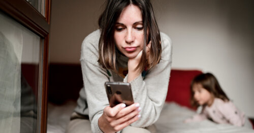 woman work online on mobile with toddler kid at home. tired woman sitting on the bad in a bedroom with smartphone in her hands and trying to work.